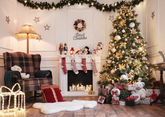 Christmassy living room with arm chair next to the fireplace full of candels. Mantelpiece covered in Christmas ornaments and toys, and stockings hanging over the fire place. Large decorated Christmas tree covered in lights and surrounded by presents